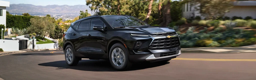 The image shows a black Chevrolet Blazer SUV driving on a road surrounded by greenery and modern homes.
